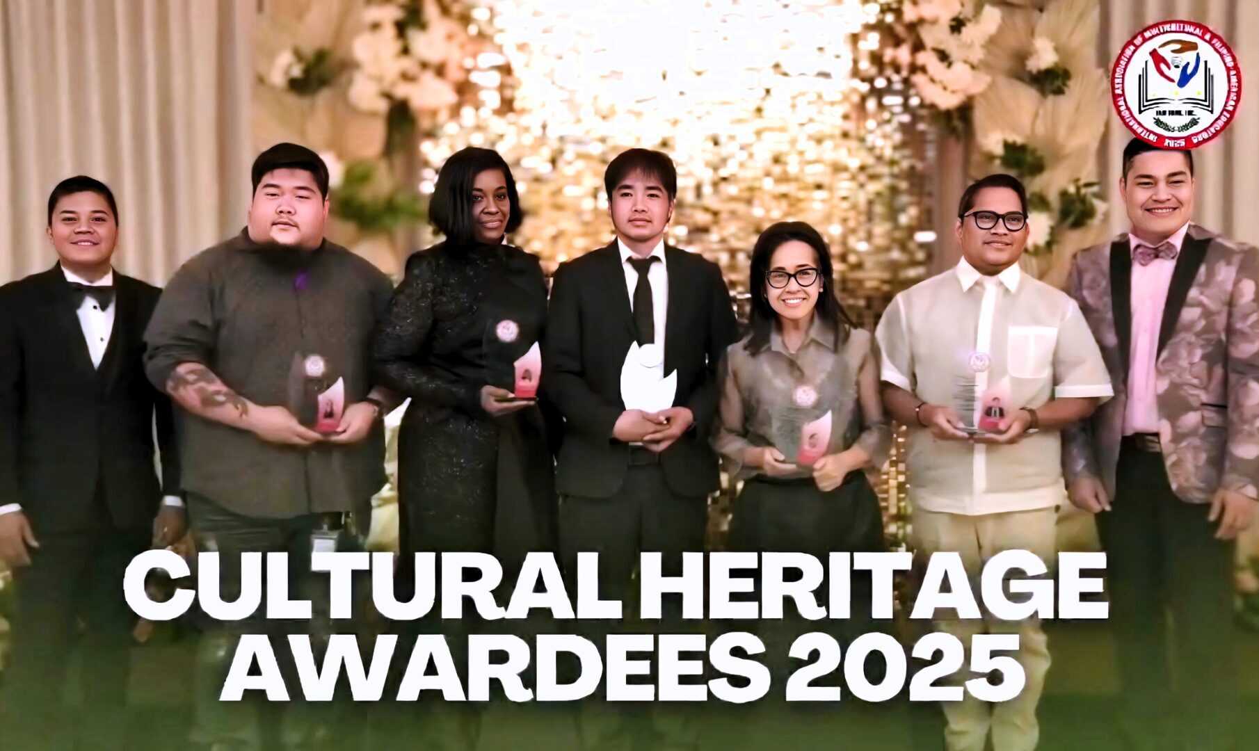 Group photo of the 2025 TEACH Cultural Heritage Awardees standing on a decorated stage, each holding an award trophy, with a warm floral and illuminated backdrop and the IAM FAME INC. logo displayed.